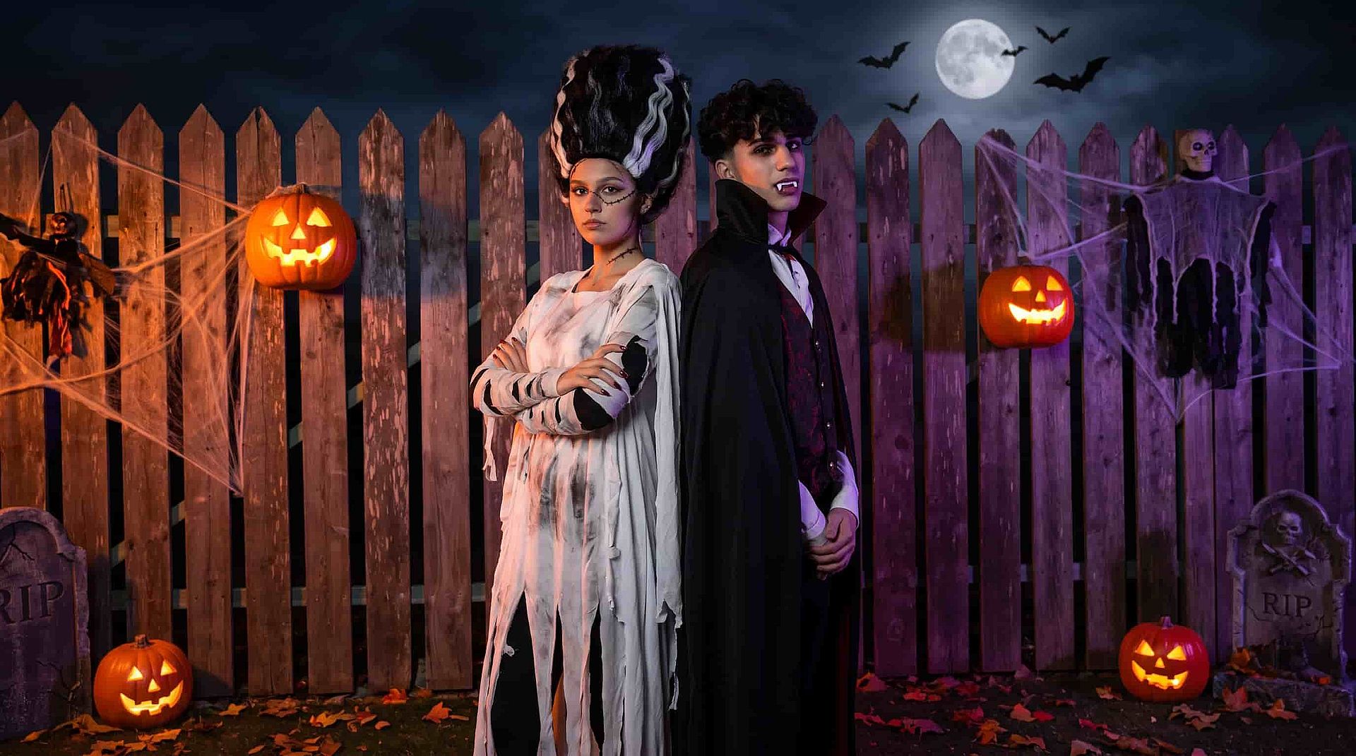 Two people in Halloween costumes posing back to back beside a wooden fence decorated with glowing pumpkins and cobwebs