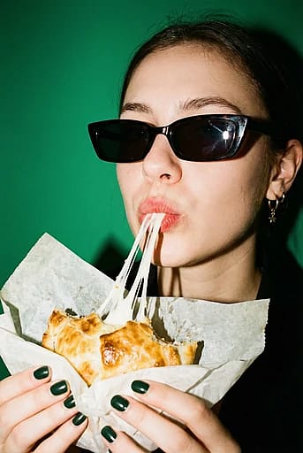 A candid high-contrast fashion portrait with harsh on-camera flash and 35mm film grain — a cheese pull from a golden baked empanada.
