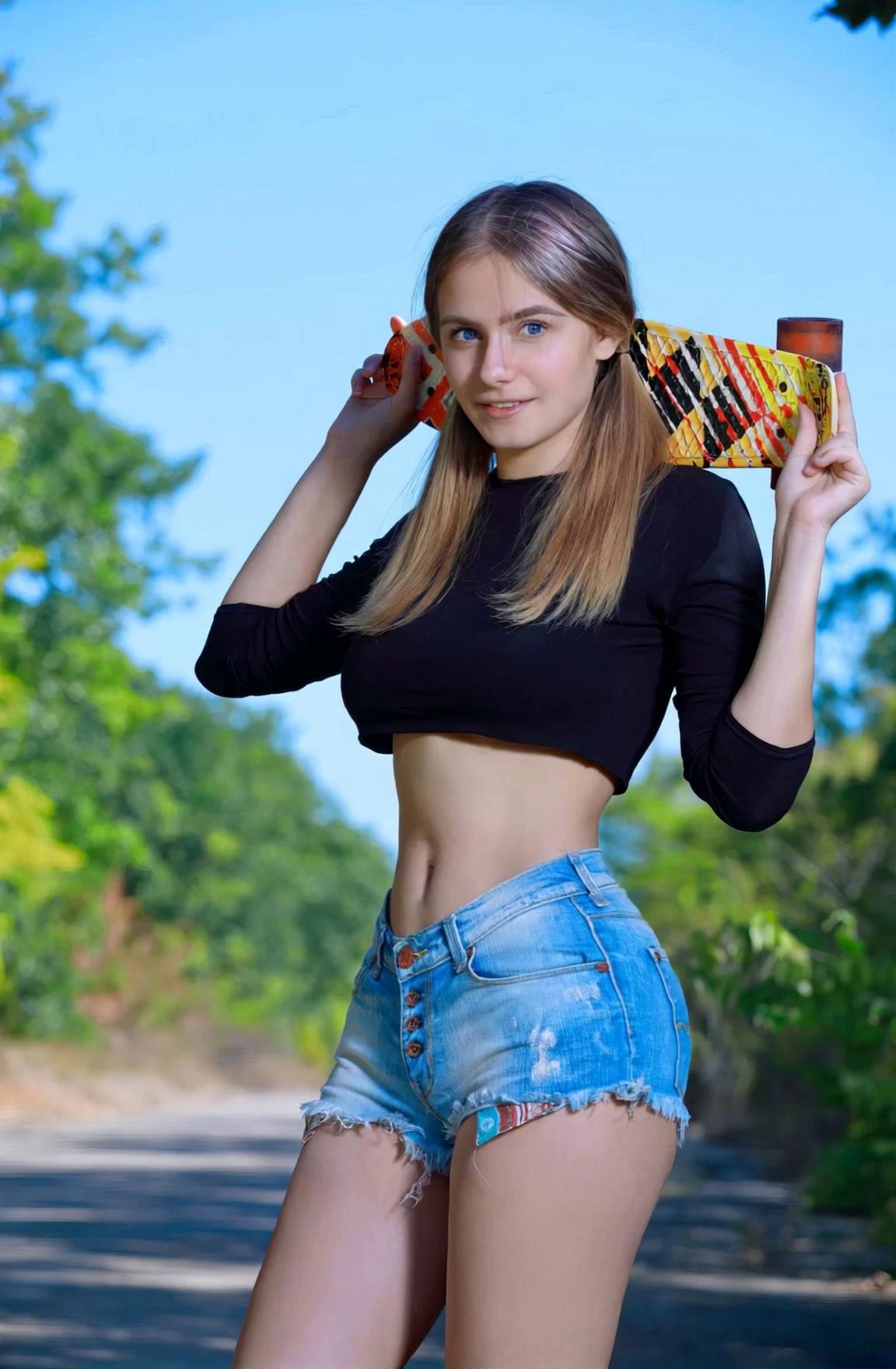 Prompt: With a bright sky in the background and vibrant greenery flanking the scene, a young woman confidently poses on a sunlit path, exuding a playful energy. She wears a fitted black crop top that accentuates her slim waist and décolletage, paired with stylish denim shorts that highlight her legs. Holding a colorful skateboard above her shoulders, she beams a smile with carefree delight, her long hair cascading down in soft waves. The girl smiles at the viewer, turns and walks away, still holding her skateboard.