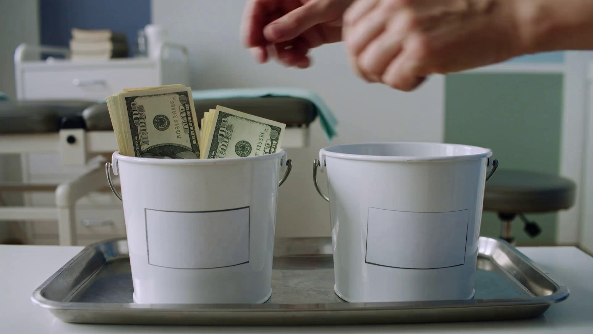 Prompt:  close-up scene where two buckets are placed on a tray in a doctor's office exam room. One bucket is initially empty and the other bucket has dollar bills in it. Both buckets have a blank label on the front. Hands move the money from the bucket with money over into the bucket without money filling it gradually. The background features medical equipment and a white wall with blue and green accents, incorporating the colors #00539B and #407E99. The lighting is natural, highlighting the contrast between the dollar bills and the exam room setting. The camera remains steady, focusing on the transfer of money, capturing the details of the bills and the movement of the hands. The video has a straightforward, instructional style, emphasizing the transfer of funds in a clean and organized manner.