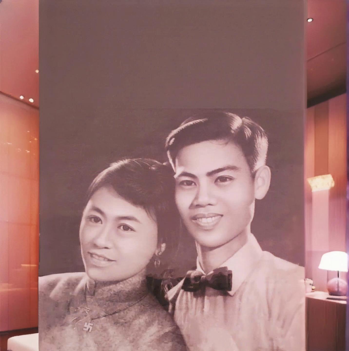 Prompt: Cinematic 10-second video, 1960s vintage film style. A young Vietnamese couple, identical to the provided photo: the man in a light shirt with a bowtie and the woman in a classic high-collar Ao Dai. They are walking hand-in-hand down a narrow village path lined with massive, ancient banyan trees with hanging roots. Dappled sunlight filters through the leaves, creating a dreamy atmosphere. They look at each other and smile tenderly. Slow tracking shot, smooth movement, soft film grain, warm nostalgic tones. "identical to the provided photo". Chất liệu phim "35mm film grain" hoặc "1960s vintage style"