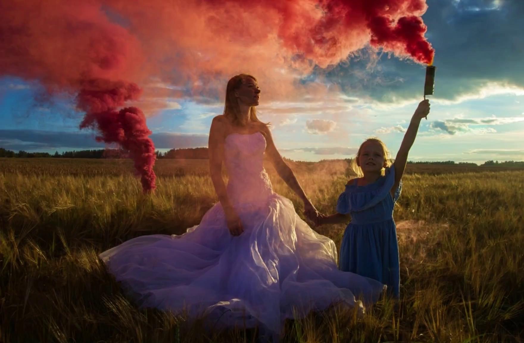 Prompt: A dreamy cinematic scene of a summer evening in a golden wheat field. A graceful woman in a flowing white wedding dress stands hand in hand with a young girl in a blue dress. They are surrounded by vibrant, swirling pink and red smoke from a flare, casting magical hues into the sky. The camera slowly circles around them as the sunlight glows warmly on the field, creating a soft, fairytale-like atmosphere. Gentle wind rustles the wheat, and the smoke dances in slow motion. The scene evokes a feeling of wonder, serenity, and timeless beauty — a blend of reality and dream.