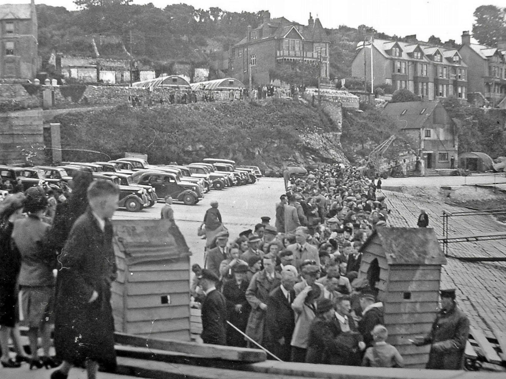 Prompt: This is a scene from the history of the town of Brixham in Devon, England.  The scene is Beakwater Beach post World war two. The people in this scene are standing in line, everyone is waiting patiently and talking, smiling and moving the way that groups of strangers do when in public. Children are more excited.Keep the full scene intact so the line of people and context remain. Avoid cropping out figures or landscape. 