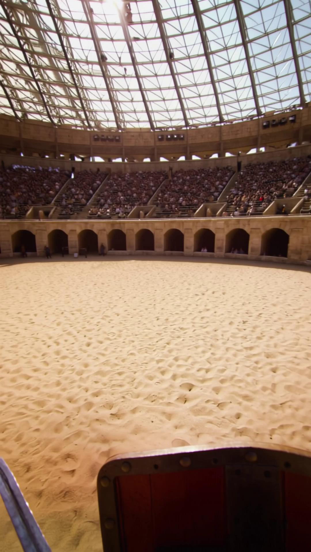 Prompt: Point of view shot, first-person perspective.
Ancient Rome, Colosseum arena, daytime.

The camera is the eyes of a gladiator standing alone on the sand-covered arena floor, at ground level.
The arena floor is completely empty except for the gladiator.
No people, no animals, no objects on the sand.

In the foreground, always visible:
	•	LEFT side of the frame: the gladiator’s shield held in the LEFT hand, close to the camera
	•	RIGHT side of the frame: the gladiator’s gladius held in the RIGHT hand, close to the camera
Orientation must remain consistent throughout the shot: LEFT = shield, RIGHT = sword.

The gladiator slowly turns his head to the RIGHT.
The camera pans right, revealing rows of spectators seated on the stone stands, high above the arena floor.
The crowd is far away, elevated, surrounding the arena from above.
No spectators are on the ground.

The gladiator then turns his head to the LEFT.
The camera pans left, revealing more spectators on the opposite side, same scale, same distance, same elevation.

Finally, the gladiator faces forward, toward the center.

In the distance, at the far side of the Colosseum, above the stands, is the imperial box (pulvinar).
The emperor is seated there, small and distant, human-sized, not enlarged.
The emperor appears far away, framed by architecture, never close to the camera, never oversized, never dominating the frame.

The emperor’s scale must match real human proportions relative to the Colosseum’s size.
He is clearly elevated, but not gigantic.

The gladiator then raises the gladius upward with the RIGHT hand, pointing it toward the imperial box.
The LEFT arm keeps the shield lowered and steady.

The crowd roars from all sides above.
The arena floor remains empty.

No camera zoom toward the emperor.
No distortion of scale.
No giant figures.

Cinematic realism, strict perspective accuracy, realistic human scale, immersive POV, dramatic sunlight, historical accuracy.
