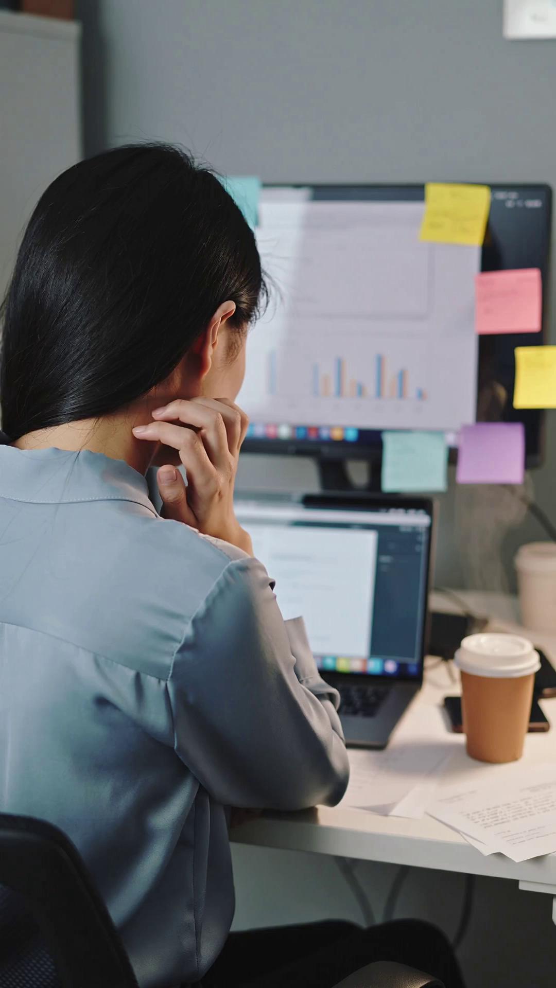Prompt: A young Asian woman in her mid-20s sitting at a modern office desk, shown from behind and slightly to the side so her face is not visible. She is in a small, cluttered workspace with a laptop open, multiple browser tabs and charts on the screen, sticky notes around the monitor, and papers scattered on the desk. Her shoulders are tense and slightly hunched, one hand rubbing the back of her neck, the other resting on the keyboard. Soft, cool office lighting, late evening mood, a half-finished coffee cup beside the laptop. The scene should clearly show work stress and overwhelm through body language and environment, but her face is completely out of frame. Cinematic, realistic, 9:16 vertical aspect ratio, shallow depth of field.