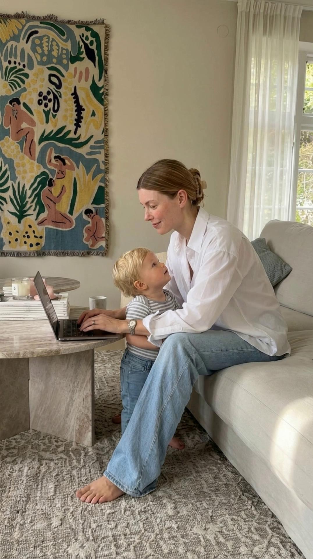 Prompt: The same young woman from the reference photo: fair cool-toned skin, soft rosy cheeks, faint freckles, blue eyes, and straight light-brown hair parted in the middle and clipped back casually. Her facial features, colouring, and proportions match her exactly.
She is sitting on a soft neutral sofa in the same living-room setting as the inspiration image: warm textured rug, stone coffee table with books, sheer curtains filtering natural daylight, and a colourful woven tapestry hanging on the wall.

She wears the same outfit style as in the inspiration photo — an oversized white button-up shirt with relaxed light-wash jeans, comfortable and casual, barefoot.
The toddler is the same baby from the reference image but at toddler age: same soft light hair, same round face, same fair skin tone. The toddler wears a striped t-shirt and light-wash toddler jeans.

Scene action:
The mum sits comfortably with her laptop open on her lap, typing gently with a focused but relaxed expression. After a few seconds, the toddler toddles over and wraps their arms around the mum’s waist. The mum looks down with a soft smile, pauses her typing, and lifts one arm to embrace the toddler.
The toddler then leans their head against the mum’s chest, holding on affectionately. The mum rests her cheek briefly on the toddler’s head, smiling warmly. Soft, realistic motion; natural toddler movements; tender mother–child moment.

Lighting is soft and warm, with natural daylight streaming through sheer curtains. The mood is calm, nurturing, and beautifully aesthetic, with smooth camera motion or static framing like a cozy lifestyle clip.
