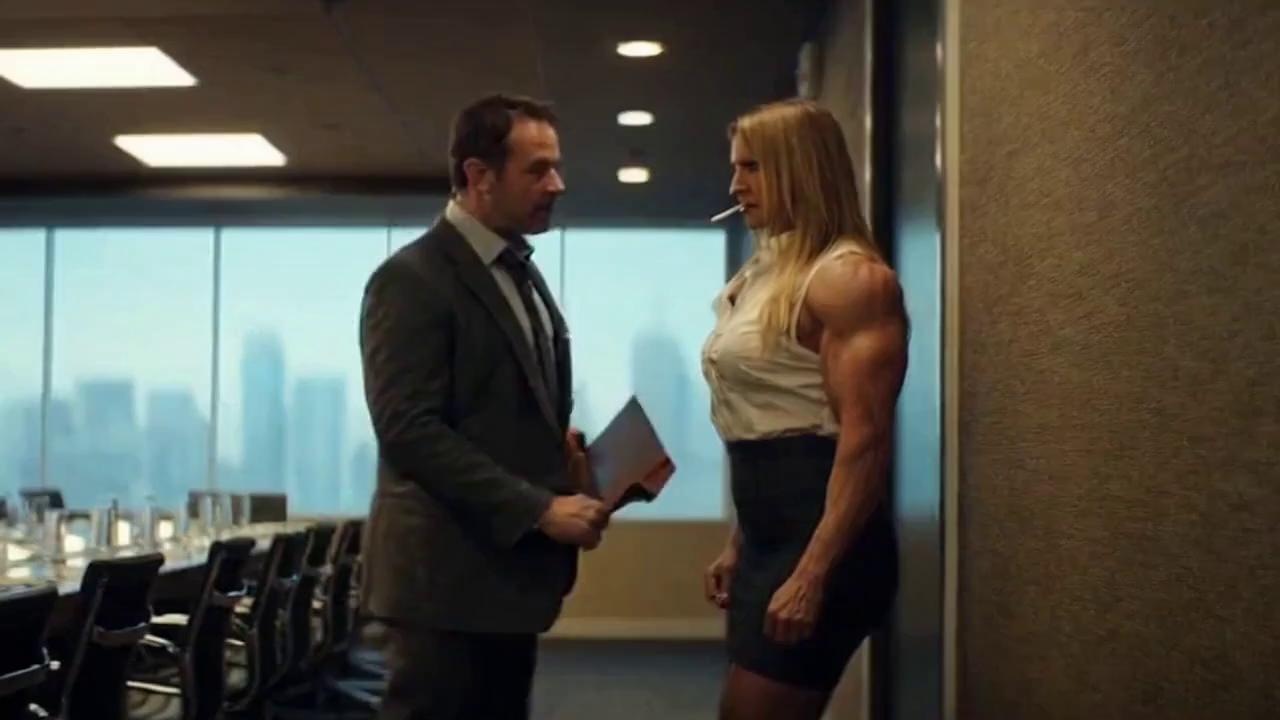 Prompt: In a cinematic scene in an action/drama movie, a muscular business woman deals with a rude male colleague violently. Grace is a tall beautiful muscular female bodybuilder with a steroid-enhanced physique, long blonde hair, dark red lips and long red fingernails wearing a dark business skirt, high heels, a sleeveless low-cut dress shirt stretched tight by her huge chest and exposing her big biceps. Bob is her colleague, dressed in a gray suit and loosened tie. Grace and Bob just finished a business lunch alone in the conference room. Grace is leaning against the wall, a cigarette dangling from her lips. While he walks past her, Bob says “Nice rack, honey!” Grace angrily grabs Bob by his jacket and slams him hard against the wall, lifting him until his feet are dangling off the floor. Grace says “You want to apologize for your rude behavior, Bobby-boy?”, her cigarette smoke causing Bob to cough. Grace persuades Bob to call her “ma’am” in his choked, stammering response.