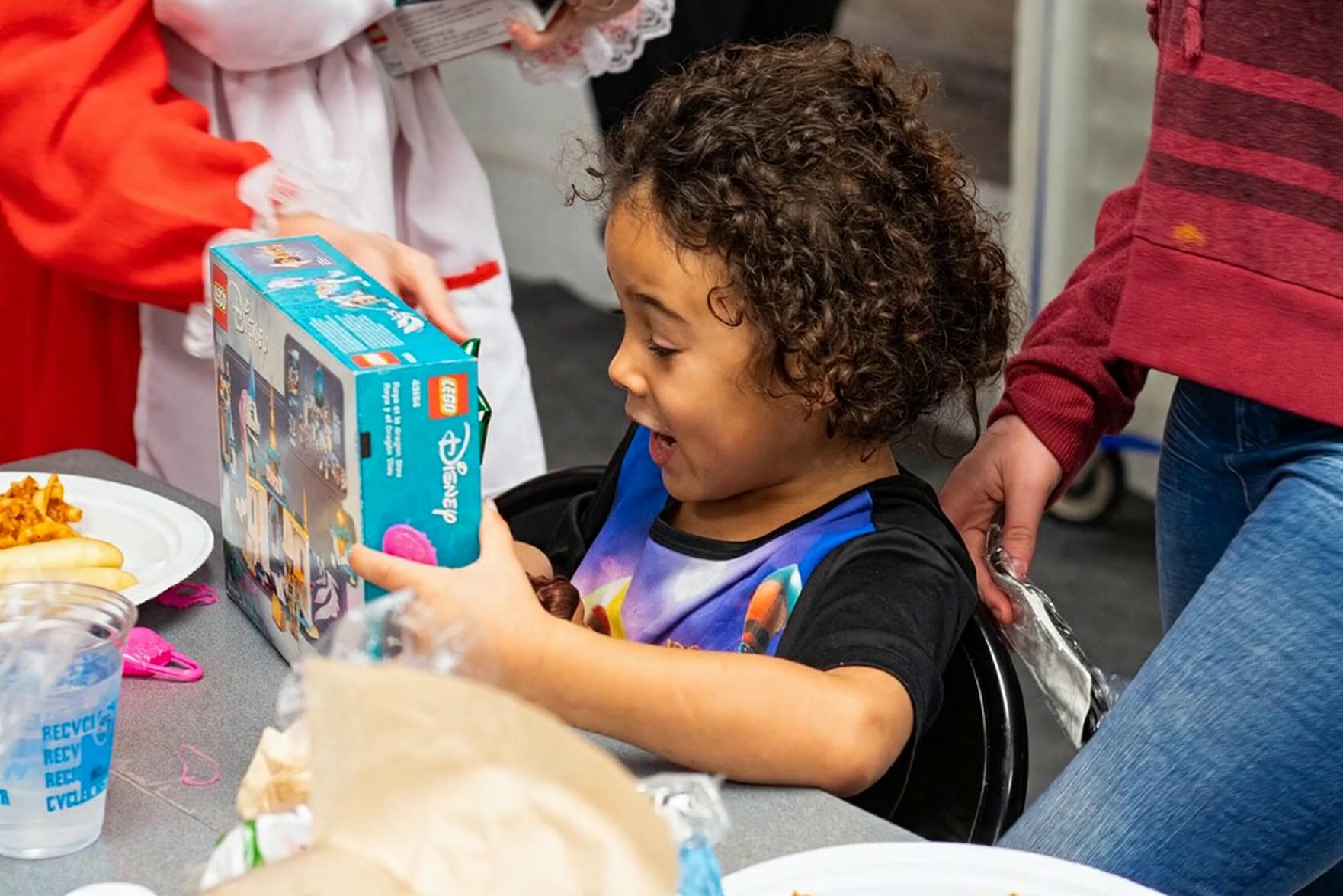 Prompt: “Create a short, realistic video from the provided image. The young girl remains exactly the same in appearance—same face, hair, skin tone, clothing, and body proportions. The environment, table, food, Lego box, and people around her remain unchanged.

The girl becomes visibly excited as she holds the Lego box: her eyes widen, her mouth opens into a joyful smile, and her body leans forward slightly with enthusiasm. She makes small, natural movements—her hands shift on the Lego box and her shoulders lift with excitement.

After a moment, she turns her head and looks up toward the woman in the red and white dress beside her, smiling excitedly as if reacting with gratitude or surprise. Her expression clearly communicates joy and excitement without exaggeration.

Camera remains stable with no zoom or angle change. Lighting stays consistent and natural. Motion is subtle, realistic, and smooth. No changes to facial structure, hair, clothing, background objects, or other people.”