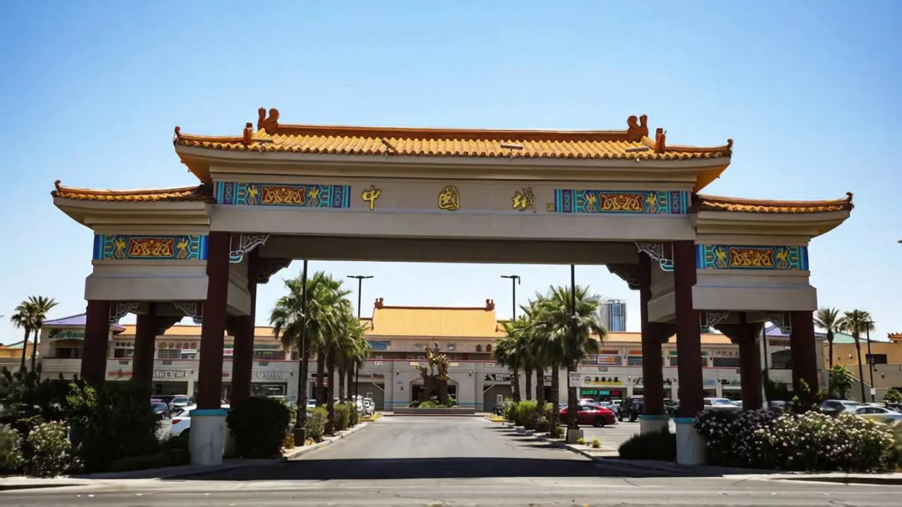 Prompt: A cinematic, slow dolly push-in shot toward a grand Chinese-style archway entrance to a shopping plaza, inspired by Las Vegas Chinatown. The ornate gateway has bold red columns, golden roof tiles, and colorful trim with traditional patterns and Chinese characters. The lighting is dramatic and moody—set at dusk with a soft purple-orange sky casting warm highlights and deep shadows across the scene. Palm trees frame the pathway leading into the plaza, where storefronts and a statue are visible in the distance. The camera glides forward slowly and steadily, capturing the depth and detail of the architecture. Ambient lantern-style lighting and glowing neon signs begin to turn on as the evening sets in. The atmosphere is calm, cinematic, and slightly mysterious.