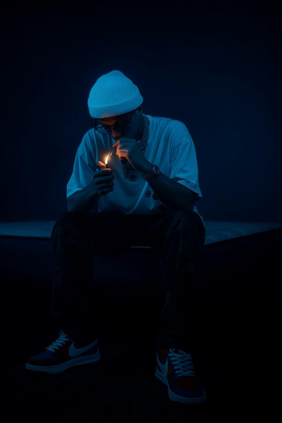 Prompt: A cinematic nighttime scene based on a still photo. A young man sits in a dark, minimal environment under deep blue lighting. He wears a white knit beanie, oversized light-colored t-shirt, dark pants, and streetwear sneakers. He is seated, leaning forward, holding a lighter close to his face.

At the start, everything is almost completely still, except for the small orange flame flickering softly, illuminating his hands and lower face. After a few seconds, he slowly inhales, slightly lifting his head without fully revealing his face. The flame briefly brightens, creating a strong contrast between warm orange light and cold blue tones.

Near the end, he exhales smoke downward and forward in slow motion. The smoke blends into the dark blue background as the camera slowly pushes in (subtle dolly-in). The flame goes out, fading into darkness. The mood is introspective, calm, and focused. Cinematic, realistic, low-key lighting, shallow depth of field, film grain, smooth motion, 24fps, high quality, music video promo aesthetic.

Negative prompt: cartoon, anime, illustration, low quality, blurry, overexposed, oversaturated, fast motion, shaky camera, distorted body, extra limbs, bad anatomy, visible face details, daylight, busy background

