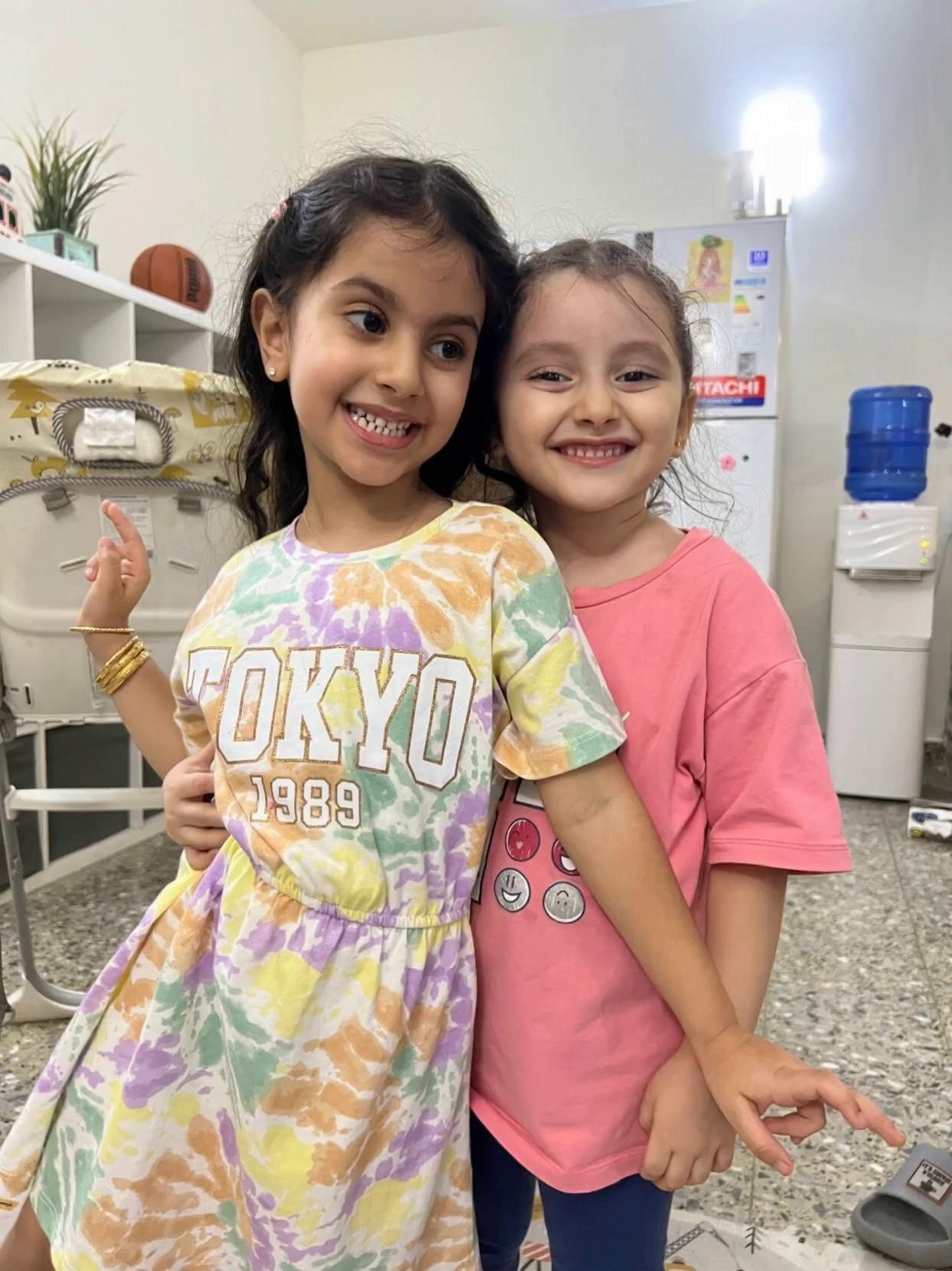 Prompt: Two little girls dancing happily together in a cozy living room. They hold hands, spin, and laugh with joy. Then they perform a small playful flip move together, smiling brightly. The lighting is warm and natural, giving a cheerful home atmosphere.