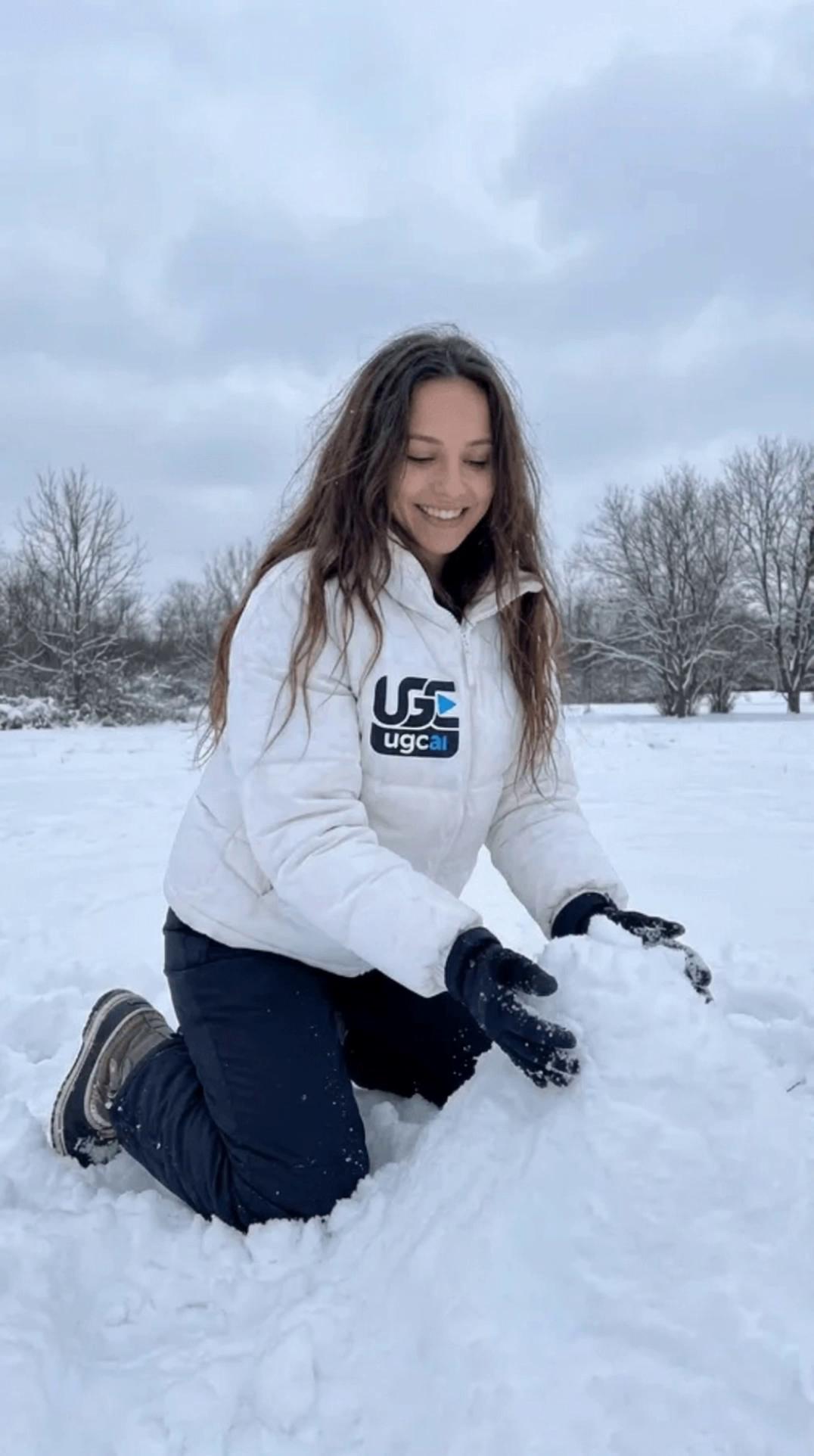 Prompt: Cinematic natural realism, full-frame 35mm anamorphic, soft bloom, subtle film grain, shallow depth of field, natural overcast lighting, neutral color grade, 24fps with natural motion blur. Stabilized handheld gimbal: start medium shot and do a slow gentle dolly-in with a slight left-to-right arc, minimal organic micro-jitter. Keep focus primarily on her face with a brief, subtle focus attention shift toward her hands (very slight, not dramatic). She moves naturally (real-time): warm genuine smile that turns into a small quiet laugh, eyes mostly looking down while she lightly pats and shapes with her hands, then a quick glance off-camera to the side and back down, natural blinking and a tiny head tilt. Background stays calm with faint breeze movement and subtle drifting particles; soft atmospheric warmth, candid and approachable.