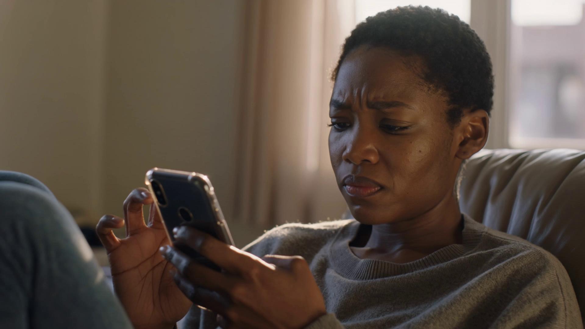 Prompt: lAfrican woman in her early 30s sitting alone in a modest, softly lit apartment in a U.S. city, evening light coming through a window, she scrolls through her phone with a tense expression, then pauses as she reads a message, her face slowly softens into visible relief, subtle exhale, natural skin texture, handheld camera, cinematic lighting, shallow depth of field, realistic environment, not staged