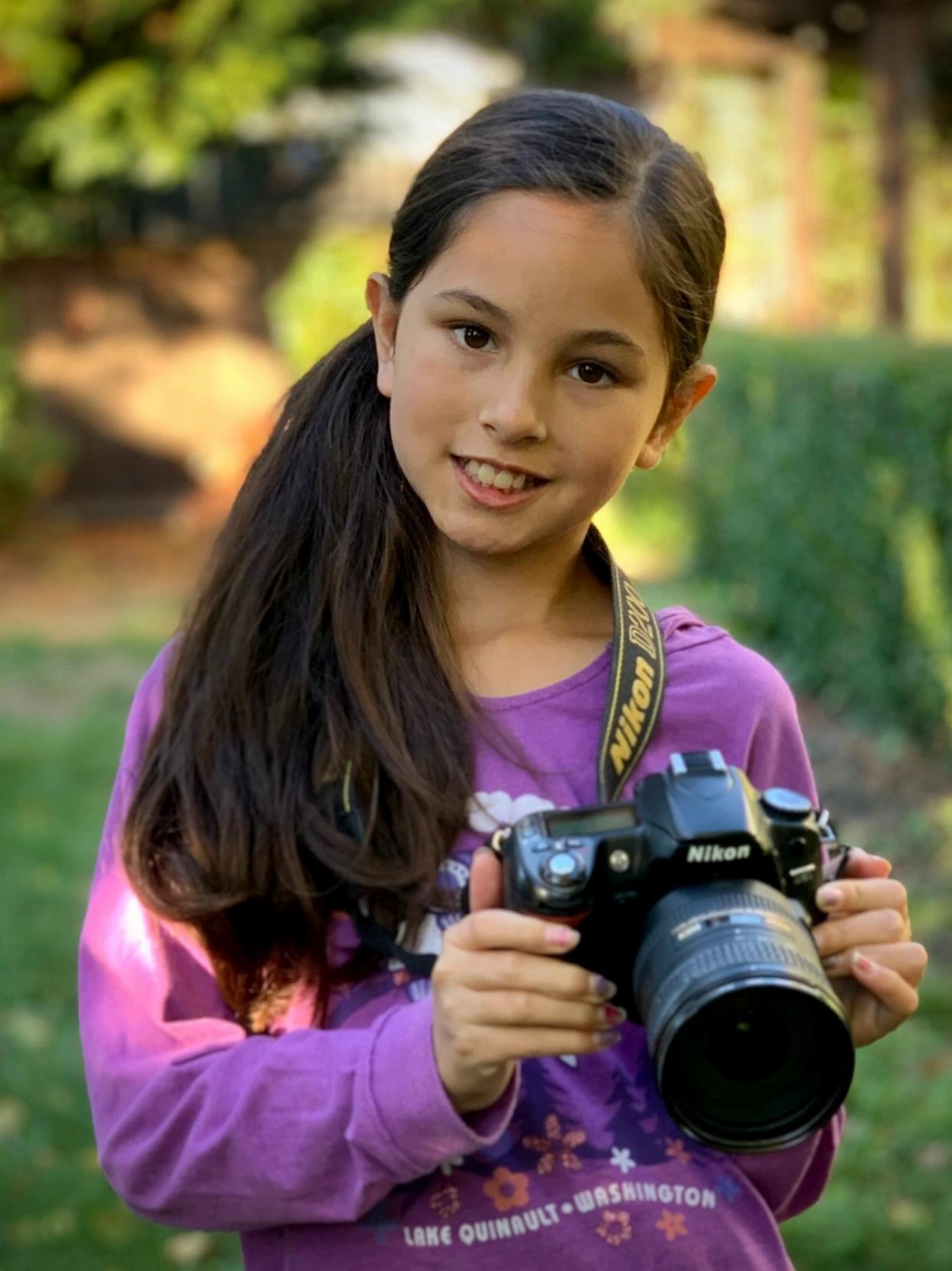 Prompt: Have the girl bring the camera up to her eye, take a picture and put it back down and then strike a pose with her hand on her hip.