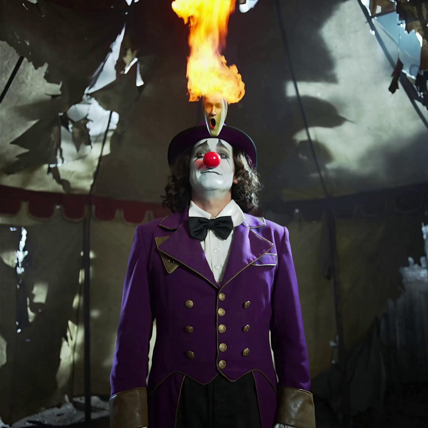 Prompt: Inside a tattered, flickering circus tent, the clown stands center ring. He breathes fire into the air — the flames twist into screaming faces that fill the tent, reflecting off the torn canvas walls. The tent flaps violently in the wind as shadows of unseen spectators stretch along the walls. The clown claps, and the fire reshapes into glowing balloons that float toward the tent ceiling. He lazily hands one to the camera. It pops — sending sparks that burn bright against the striped interior — and the screen cuts to black.