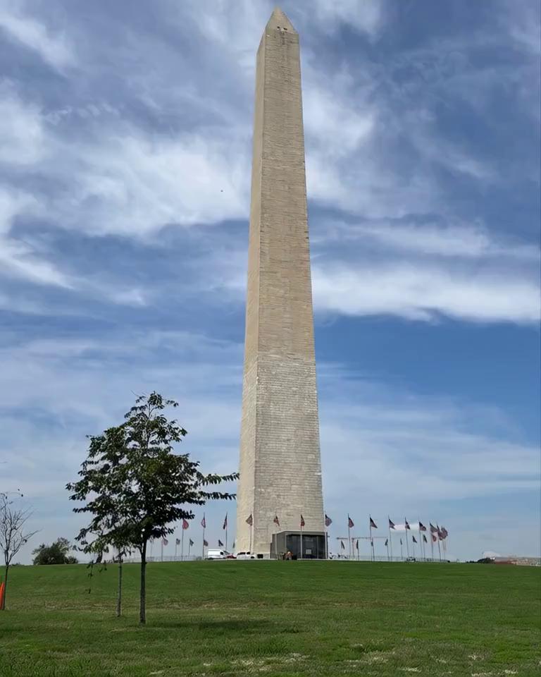 Prompt: yper-realistic, 8K, 16:9 format, cinematic time-lapse scene of the Washington Monument (obelisk) captured from a fixed frontal viewpoint. The sequence shows the transition from day to night: dynamic movement of clouds drifting across the sky as daylight gradually fades. The sky shifts through warm golden sunset tones into a deep, starry night. Finally, a large full moon rises, casting a soft natural glow and creating a subtle moonlight reflection on the stone surface of the obelisk, gently illuminating the monument and the surrounding grassy area. A gentle nocturnal breeze flows through the scene, adding an atmospheric and immersive touch to the night. The breeze causes the American flags encircling the monument to wave softly and naturally. The transitions of light, shadow, sky movement, and flag motion should feel smooth and cinematic, emphasizing the monument’s height and the serene yet majestic atmosphere of the changing sky. Ultra-detailed hyper-realistic textures for the stone, grass, flags, and sky, with seamless lighting transitions.