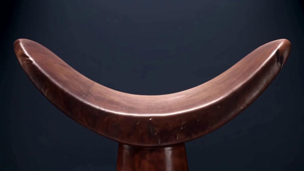 Prompt: this afar pillow is it hold head like pillow  this pillow design like shoulder cross 
Storyboard: Pillow as Living Anatomy
Scene 1 – Introduction (3–5 sec)

Visual: A traditional wooden headrest floats in darkness. Soft spotlight above.

Camera: Slow zoom-in from front, orbiting slightly to the side.

Mood: Sacred, mysterious, like a relic.

Scene 2 – First Transformation (5–7 sec)

Visual: The curved top of the headrest begins to glow faintly.

Action: The curve stretches, morphing into the rounded collarbone and shoulder line of a skeleton.

Camera: Tracking shot from side, moving closer to highlight the morph.

Effect: Smooth morphing, like wood turning into bone.

Scene 3 – Spine Connection (4–6 sec)

Visual: The central stem of the pillow elongates upward into a neck vertebrae.

Action: As it extends, faint glowing lines travel down it, symbolizing energy.

Camera: Tilt upward to follow the stem transforming into a spine.

Effect: Organic growth, spine forming vertebra by vertebra.

Scene 4 – Ribcage Expansion (6–8 sec)

Visual: The wide base of the pillow spreads out, morphing into a ribcage.

Action: The ribcage expands and contracts slowly, showing breathing movement.

Camera: Slow push-in from front, chest filling frame.

Effect: Breathing rhythm synced with sound (inhale/exhale).

Scene 5 – Overlay Moment (5–7 sec)

Visual: Half wood / half skeleton overlay. The pillow and human form flicker and blend together.

Action: Transition back and forth — pillow → skeleton → pillow.

Camera: Static, centered, focusing on the morph.

Effect: Dreamlike dissolve, glowing edges.

Scene 6 – Return to Object (3–5 sec)

Visual: The skeleton fades away, leaving only the wooden pillow again.

Action: Pillow rests calmly on a flat surface.

Camera: Pull-back shot, wide angle.

Mood: Peaceful, grounded, like the pillow carries the memory of the body. 