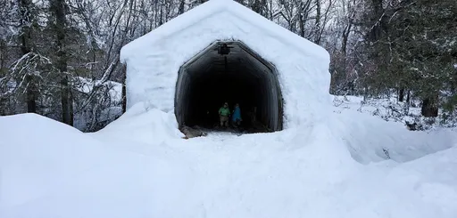 Prompt: In Alberta, Canada, winter arrives with a blanket of snow so deep, locals are compelled to dig tunnels for barn access. Amidst the  snow a farmhouse stands resilient. A tunnel, laboriously carved through towering snowdrifts, guides the way to the barn. Inside, farmers tend to the animals, their breath visible in the chilly air showing the spirit of rural Canadian life