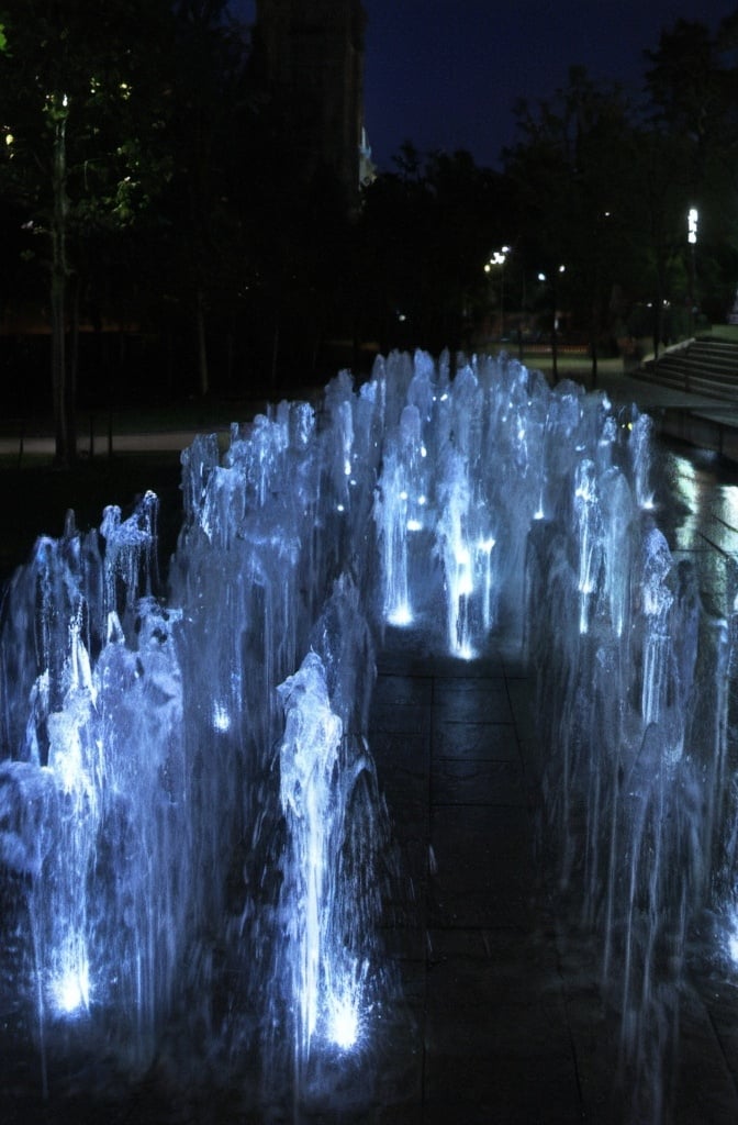 Prompt: a large fountain of water with lights on it's sides at night time in a park area.   kinetic art, volumetric lights, an abstract sculpture