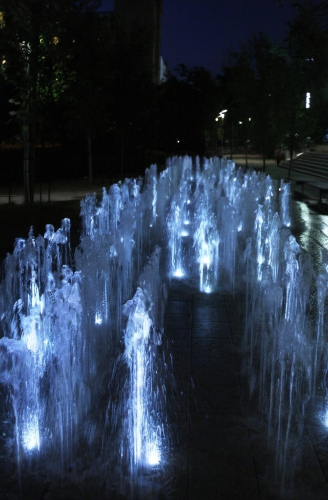 Prompt: a large fountain of water with lights on it's sides at night time in a park area. Steven M. Tilley,  kinetic art, volumetric lights, an abstract sculpture