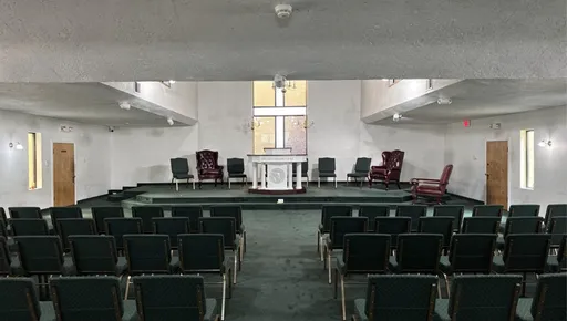 Prompt: Redesign this church. Remove all existing furniture and add all new green pattern chairs in the audience and 4 to the pulpit. Add  2 leather chairs to the pulpit. Add a new podium in the pulpit. Add a white communion table in the center of the church, near the front. Leave the 2 office doors on the right and left at the front of the church