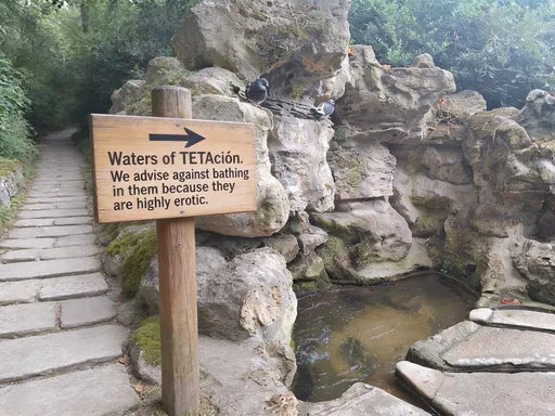 Prompt: Sobre el sendere, un crtrl de madera sobre un poste indica una flecha hacia el agua diciendo "Aguas de la TETAciòn. Desavonsejamos bañarse en ellas porque son altamente erotizantes"