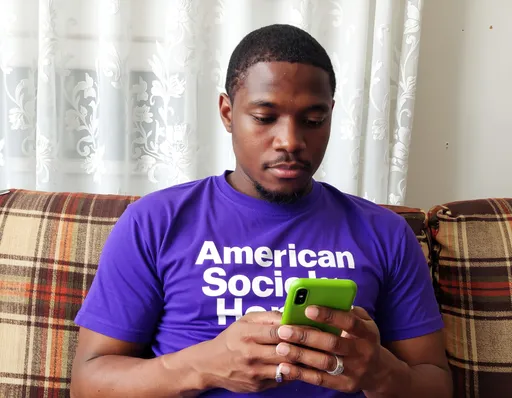 Prompt: This photograph depicts a young Black man with a medium-dark skin tone, short-cropped black hair, and a clean-shaven face. He is sitting on a plaid-patterned couch with brown and orange diagonal lines. The man is wearing a vibrant purple t-shirt with white text that reads "American Society Hope." He is focused intently on a green smartphone he is holding with both hands. His right hand, which is adorned with a silver ring on the ring finger, is positioned above the phone, while his left hand is below it. The background features a white, floral-patterned curtain that adds a subtle texture to the scene. The lighting is soft and natural, highlighting the man's face and the colors of his shirt and phone. The overall mood of the photograph is calm and contemplative, with the man's concentrated expression suggesting he is deeply engaged with the content on his phone. The image is sharply focused, capturing the details of the man's t-shirt, the couch's texture, and the floral pattern of the curtain.
