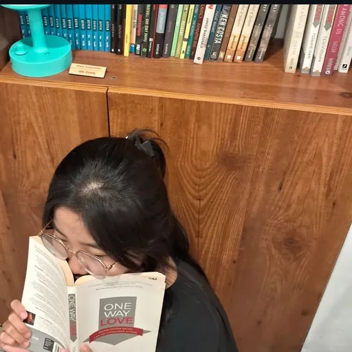 Prompt: Nestled against a warm wooden backdrop, a young woman with dark hair tied in a loose ponytail sits quietly, her face partially concealed behind an open book titled "One Way Love." Her round glasses reflect a sense of contemplation as she gazes at the pages, seemingly immersed in the narrative, embodying a moment of peaceful introspection. Surrounding her, a neatly arranged bookshelf showcases a collection of colorful books in various genres, complemented by a whimsical turquoise lamp that adds a pop of color to the serene setting. The overall atmosphere conveys a cozy, intimate space perfect for reading, capturing a mood of curiosity and solitude.