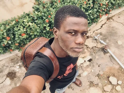 Prompt: This photograph depicts a young African-American man standing outdoors against a weathered, beige concrete wall adorned with green foliage and small, red flowers. He is looking directly at the camera with a neutral expression, his dark brown eyes slightly narrowed. He has a close-cropped, short haircut and a light stubble on his face. He is wearing a black t-shirt with a red graphic design on the chest and light blue jeans that are slightly worn. Over his right shoulder, he carries a brown leather backpack with a textured pattern and a single strap that rests on his back. The ground beneath him is cracked and uneven, with patches of dirt and small rocks. His brown leather sandals are visible at the bottom of the frame. The photograph is taken from an elevated angle, emphasizing his facial features and upper body. The overall atmosphere is casual and urban, with a focus on natural light and everyday elements.