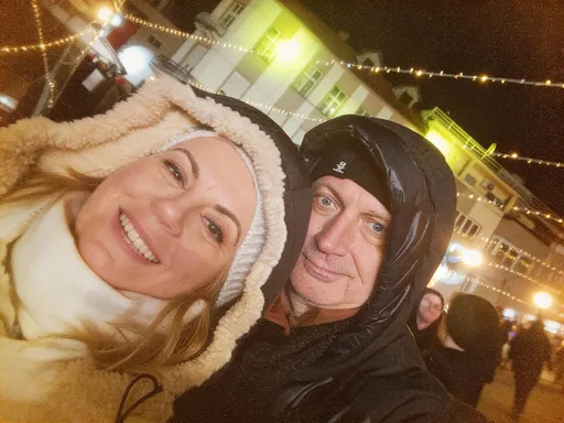 Prompt: This photograph captures a close-up of a smiling middle-aged Caucasian couple standing outdoors at night. The woman, positioned on the left, has fair skin, light brown hair, and is wearing a white, fluffy, knitted beanie with ear flaps, and a cream-colored turtleneck. She has light blue eyes and is smiling broadly, showing her teeth. The man, on the right, has a slightly weathered face with wrinkles and light blue eyes. He is wearing a black, glossy, puffer jacket and a black, hooded beanie. His expression is more neutral but he is also smiling slightly. The background features a dimly lit urban setting with string lights crisscrossing above, creating a festive atmosphere. There are blurred lights from street lamps and possibly other people in the background. A white building with green-lit windows is partially visible behind them. The overall image has a warm, slightly grainy texture, typical of nighttime outdoor photography. The couple's close proximity and the festive string lights suggest they might be enjoying a holiday or winter event.