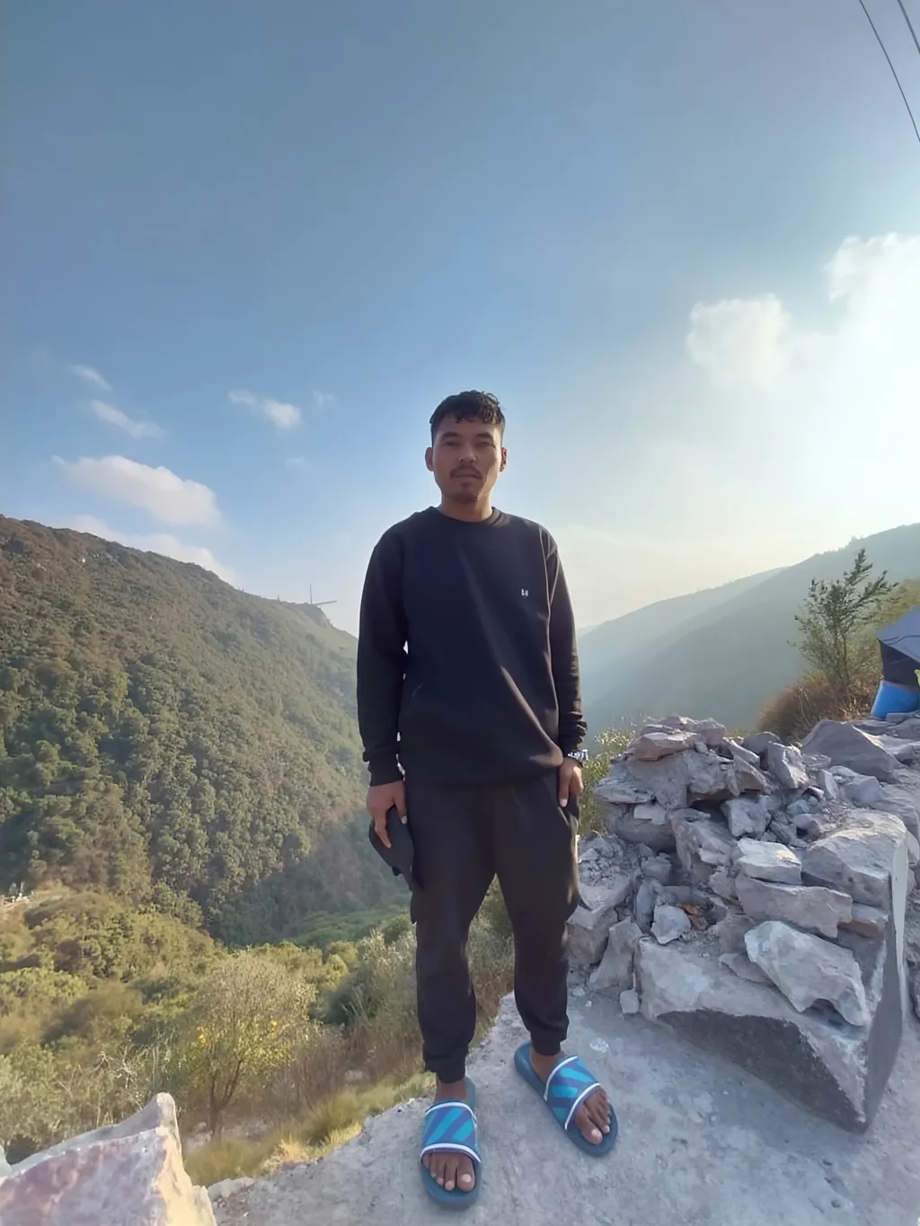 Prompt: Standing against a backdrop of majestic hills under a clear blue sky, a young man exudes a calm yet contemplative demeanor. Dressed in a black sweatshirt and matching sweatpants, his casual look is complemented by blue flip-flops, which contrast with the rugged terrain around him. As he holds a device in one hand, the rocky outcrop beside him adds a touch of natural ruggedness to the scene, while the lush greenery in the distance suggests a sense of tranquility and connection with nature. The sunlight softly illuminates the landscape, creating a serene, peaceful atmosphere that invites reflection on the beauty of the outdoors.