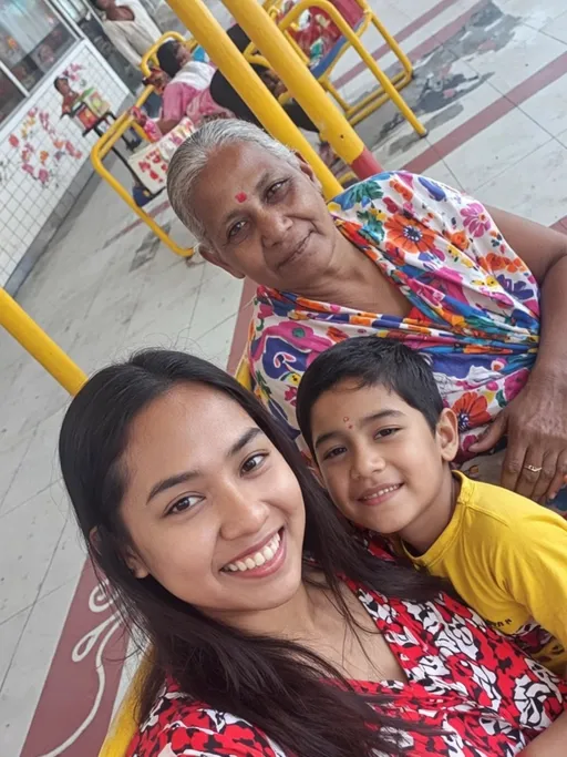 Prompt: A warm scene unfolds as three generations of women share a moment together, seated on a vibrant yellow swing set in a public space. The foreground captures a young woman with long, dark hair and a patterned red and white outfit, smiling brightly beside a little girl clad in a cheerful yellow shirt, both exuding playful camaraderie. In the background, an elderly woman with a gentle smile and greying hair, draped in a colorful sari, looks on with pride and affection. Soft, natural light illuminates their faces, conveying a sense of joy and familial bond, while the floor beneath them showcases a playful design that complements the lively colors, enhancing the overall warmth of this intimate gathering.