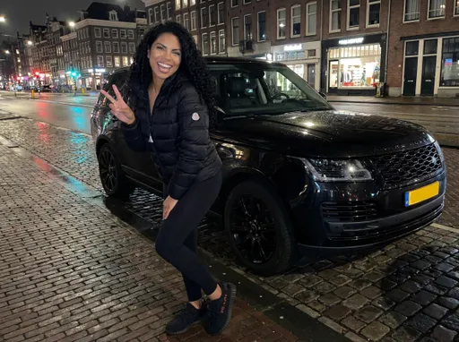Prompt: The image shows a photo of the same woman standing on a cobblestone street somewhere in the Netherlands at night. She is wearing a black Moncler jacket, black leggings, and black Yeezy sneakers . She has curly dark hair and is smiling at the camera. In the background, there is a black Range Rover parked next to her. The woman is making a peace sign with her hand and appears to be posing for the photo.