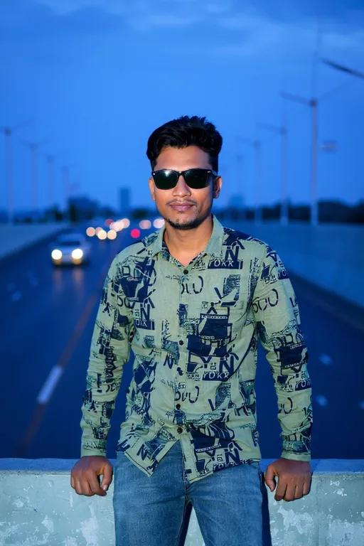 Prompt: Photograph of a young South Asian man with a medium build and short, styled black hair. He wears black sunglasses, a green, patterned button-up shirt with abstract designs in black, and blue jeans. He leans against a concrete barrier on a wide, empty highway with blurred traffic in the background, under a twilight sky. His expression is confident, with a slight smile. The highway is lined with wind turbines in the distance, adding a modern, industrial touch to the scene. The overall color palette is cool with blue and green tones, enhancing the evening atmosphere.