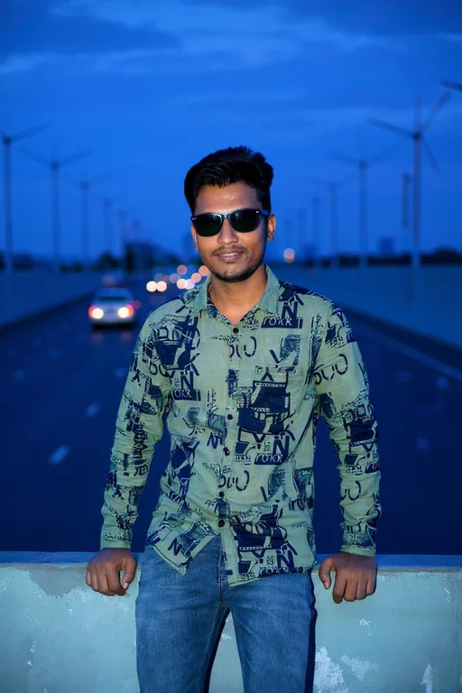 Prompt: Photograph of a young South Asian man with a medium build and short, styled black hair. He wears black sunglasses, a green, patterned button-up shirt with abstract designs in black, and blue jeans. He leans against a concrete barrier on a wide, empty highway with blurred traffic in the background, under a twilight sky. His expression is confident, with a slight smile. The highway is lined with wind turbines in the distance, adding a modern, industrial touch to the scene. The overall color palette is cool with blue and green tones, enhancing the evening atmosphere.