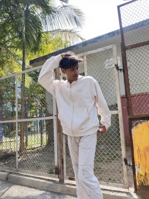 Prompt: Bathed in warm sunlight, a young man stands casually against a weathered, metal fence, his body angled in a relaxed yet confident pose. Dressed in a matching light-colored hooded jacket and trousers, he brushes his tousled hair back with one hand while the other rests on his hip, exuding a laid-back charm. Behind him, vibrant greenery with palm fronds sways gently in the breeze, contrasting with the rustic, faded colors of the background. The scene captures a tranquil moment in a sunlit outdoor space, conveying an air of youthful ease and serenity. The overall composition highlights the interplay of light and shadow, enriching the casual yet dynamic vibe of the image.