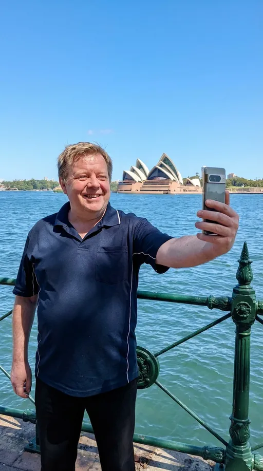 Prompt: he is standing in a background of the opera house with green cast iron fence. He is standing behind the green cast iron fence. He is taking a selfie with his google pixel pro in his right and and he is smiling  please make sure his facial and hair style is the same. Picture number two is the image of the sydney opera house with the green cast iron gates
