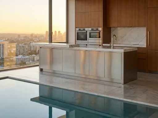Prompt: This photograph captures a modern, luxurious kitchen with a stunning urban skyline view in the background. The kitchen features a sleek, stainless steel island with a smooth, white marble countertop, positioned centrally. The island has a minimalist design with a lower cabinet section and a raised bar area. Above the island, there are two built-in ovens with a matching stainless steel finish, integrated into a wooden cabinetry unit that extends across the right side of the image. The wooden cabinets have a warm, medium-brown finish with clean lines, adding a touch of warmth to the modern aesthetic.

The backsplash behind the island is also made of white marble, creating a cohesive look with the countertop. A modern, stainless steel faucet is mounted on the right side of the island. The floor is covered with large, polished marble tiles that reflect the light, adding to the kitchen's elegance. In the foreground, there is a reflective pool or water feature that mirrors the kitchen and the cityscape.

The background showcases a panoramic view of a city skyline at sunset, with tall buildings illuminated by the setting sun, casting a warm, golden glow over the scene. 