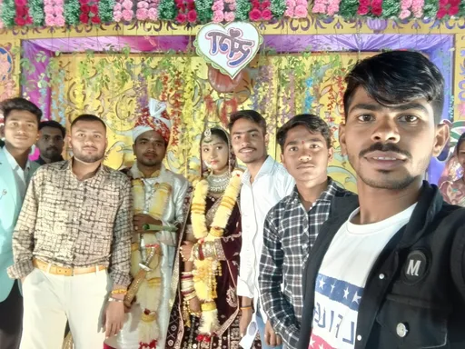 Prompt: A festive gathering unfolds in a vibrant setting adorned with intricate decorations featuring floral accents and colorful backdrops, suggesting a celebration, possibly a wedding. In the foreground, a group of young men and a woman take a selfie, their expressions filled with joy and camaraderie; the woman, dressed in traditional attire, is adorned with jewelry and floral garlands, indicative of a ceremonial role. The men, dressed in various styles ranging from casual to semi-formal, exude a lively energy, forming a striking contrast against the ornate, decorative background that hints at cultural significance. Soft lighting enhances the colors, creating a warm and inviting atmosphere that captures the spirit of togetherness and festivity. The scene conveys a sense of celebration and community, highlighting the importance of shared moments in life’s special occasions.