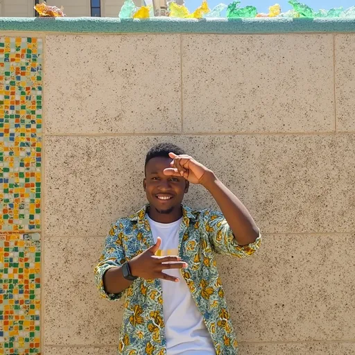 Prompt: Basking in bright sunlight, a young man poses confidently against a textured, multi-colored tiled wall, creating a vibrant backdrop. He sports a loose, patterned shirt with an eye-catching design, layering it over a simple white t-shirt, and strikes a playful pose with his hands—one forming a gun gesture while the other playfully shields his face. His expression exudes joy and charisma, with a slight smile hinting at a carefree spirit. Above him, shards of green and yellow glass crown the wall, suggesting a touch of creativity and urban edge, while the warm color palette enhances the lively atmosphere of the moment. This scene radiates youthful exuberance and a sense of fun, capturing a snapshot of life filled with energy and personality.