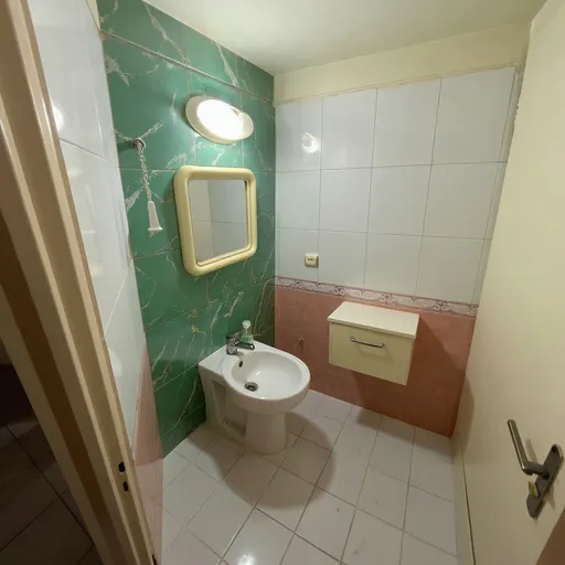Prompt: Keep the same bathroom layout, proportions, lighting, and perspective from the uploaded photo. 
Transform the bathroom to look newly renovated with a realistic, modern design. 
Use jade green marble tiles with natural veins on the wall behind the toilet. 
Use white marble tiles with elegant golden veins on the wall with the sink and mirror. 
Keep all other walls light and neutral. 
Floor tiles and door should be very light matte gray, smooth and modern. 
Install matte gold faucets, handles, and accessories. 
Add a small cream-colored vanity with a white countertop under the mirror. 
Use soft natural daylight as the lighting, creating realistic reflections and shadows. 
Maintain the same camera angle and keep all existing objects in place, just restyle the materials and colors realistically. 
Ultra-realistic photorealistic texture, 8K detail.