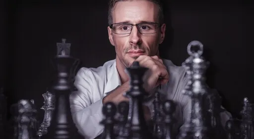 Prompt: man wearing a white button down with sleeves rolled up to elbows, and top 2 buttons undone, sitting in a oversized ornate wooden chair on opposite side of a large, elegant desk. Chess board on desk with hpandcrafted oversized glass chess pieces. He's looking forward while he rests his head on his clasped hands, which are under his chin. 

Camera angle is directly in front - 4 feet away - table height. room is dark except for the chess board and pieces that are illuminated by a single powerful light above. Style is photorealistic. Ultra detailed - 8k. Muted colors, cold, but very detailed. 