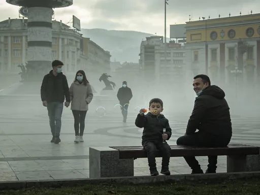 Prompt: Ultra-realistic documentary-style photograph.
Winter afternoon at Macedonia Square in Skopje, North Macedonia.
Heavy smog and fog covering the square, low visibility,
cold muted colors, grey-green tones, dull and unpolished look.

In the foreground, a father and a young child are sitting on a public bench.
The child is calmly blowing soap bubbles.
The bubbles float briefly and disappear into the fog.
The father watches the child and smiles softly, relaxed and unconcerned.
They are not wearing masks.

In the mid-ground, a young couple wearing medical face masks walk slowly
across the square, holding hands, quietly talking to each other.

In the background, a lone man wearing a medical mask walks through the fog,
partially obscured, moving away from the camera.

Monuments and buildings typical of Skopje’s main square are barely visible
through the smog, softened silhouettes, no clear landmarks.

Natural overcast lighting, flat light, no shadows.
Imperfect framing, slight atmospheric haze.
Feels like a real street photograph taken by chance, not staged.
