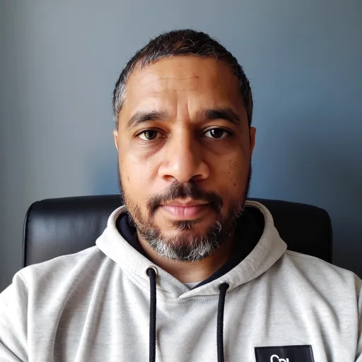Prompt: Photograph of a middle-aged Black man with a medium-dark skin tone, short black hair, and a neatly trimmed beard. He has light blue eyes and is wearing a gray hoodie with a black logo partially visible on the chest. The hoodie has a drawstring and is pulled up, exposing his head. He is sitting against a solid blue background with a black cushion behind his head. His expression is neutral, with a slight, closed-mouth smile. The lighting is even, highlighting his facial features and the texture of his hoodie. The overall composition is simple and focused on the subject's face.