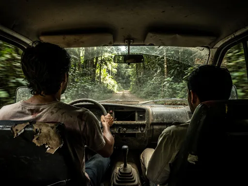 Prompt: Inside of a dirty van driving in bolivian forest. We see the driver and a passanger from behind.