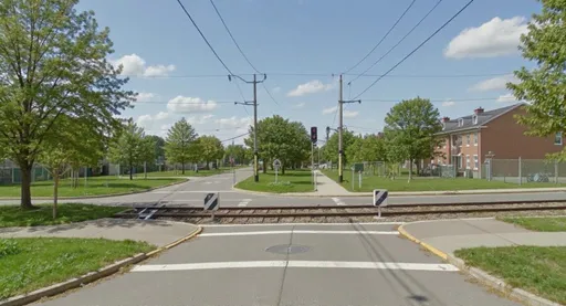 Prompt: A well-designed urban railway level crossing in a residential area, at daytime. The scene shows a fully signalized intersection with automated barriers for vehicle safety. There are clearly marked pedestrian crossings with tactile paving and robust, accessible sidewalks leading to the tracks. The road is smoothly paved, and the surroundings feature well-maintained green spaces and orderly street lighting. Emphasize safety, clear visibility, and integration into the urban fabric. Photorealistic, bright, inviting atmosphere. --ar 16:9 --v 5.2