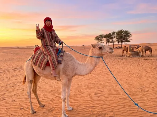 Prompt: 
A full shot shows a woman riding a camel in a sandy desert setting during sunset. She wears a red head covering, striped robe and high heels facing the camera. The camel is light colored with blue rope leading. 
In the far distance a small group of trees and a well. Around the well there are a group of camels standing.