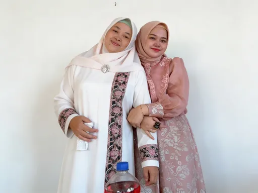 Prompt: This is a photograph of two women standing closely together, smiling at the camera. Both women are wearing traditional Malay attire, known as baju kurung. The woman on the left has a white baju kurung with intricate pink and black floral embroidery along the sides and sleeves. She also wears a white hijab with a small silver brooch at the center. Her skin is light brown, and she has a gentle smile. The woman on the right has a pink baju kurung with delicate white floral patterns and a matching pink hijab. She has a slightly darker complexion compared to the woman on the left and also smiles warmly. Her right arm is around the woman on the left, and she is holding her hand gently. She wears a black ring on her right hand. Both women have simple makeup with pink lipstick. In front of them, there is a clear plastic bottle with a blue cap containing a red liquid. The background is plain and white, drawing attention to the women and their attire. The photograph has a soft, natural light, enhancing the warm and friendly atmosphere.