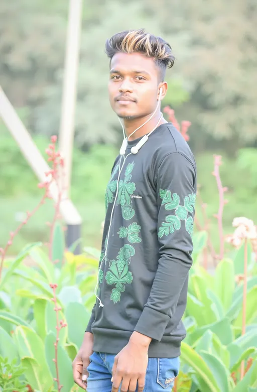 Prompt: Standing amidst lush greenery, a young man is captured with a serene expression, exuding confidence and charm. He wears a black long-sleeve shirt adorned with intricate green designs, which complements his casual blue jeans. His hair, styled with highlighted tips, adds a modern flair, while a pair of earphones hangs around his neck, suggesting a relaxed, contemporary vibe. The background features a blend of greenery and hints of blooming flowers, creating a vibrant atmosphere, possibly during daylight, enhancing the overall inviting mood of the scene.