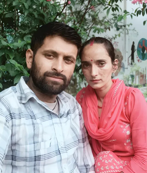 Prompt: Surrounded by lush greenery, two individuals pose closely together, capturing a moment of connection amid vibrant pink flowers that bloom behind them. The man, sporting a beard and wearing a black-and-white checked shirt, gazes directly into the camera with a neutral expression, while the woman beside him, draped in a striking red outfit, maintains a serious demeanor, her gaze slightly averted. The scene appears to be set outdoors, possibly in a garden or park, reflecting a serene atmosphere enhanced by the natural beauty surrounding them. The composition emphasizes their expressions, drawing attention to the intimate moment, while the soft lighting gently illuminates their features. Together, the image conveys a sense of companionship and tranquility against a backdrop of nature's vivid colors.